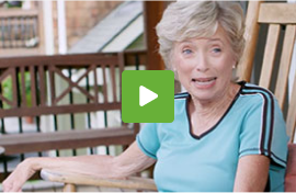 An elderly women in a light blue short-sleeved shirt, sitting on a wooden rocking chair with wooden railings and houses in the background.